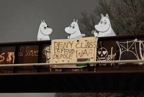 Night photo of an elevated structure with a hanging sign reading 'DENY CLASS DEFEND WAR DEPOSE,' surrounded by graffiti and three cartoon Moomin-like figures above.