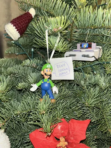 A Luigi ornament hanging on a Christmas tree beside a small toy gun and a paper tag with faint handwritten text.