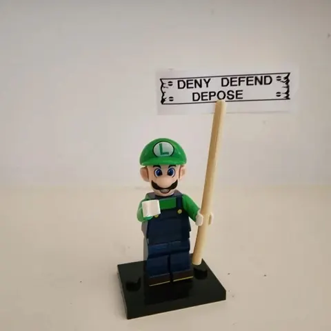 A Luigi minifigure on a display stand beneath a small sign reading 'DENY DEFEND DEPOSE.'