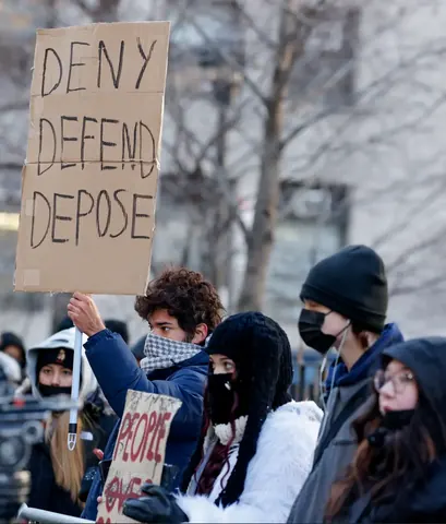 A bundled-up protester holding a cardboard sign reading 'DENY DEFEND DEPOSE' above a crowd outdoors in winter.