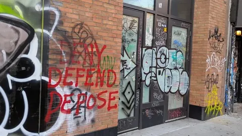 Red spray-painted 'DENY DEFEND DEPOSE' graffiti on a brick wall beside a heavily tagged doorway.