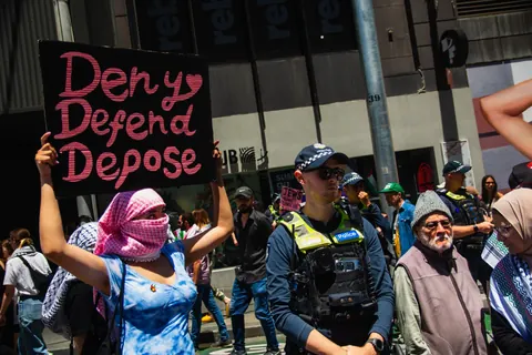 A person in a crowd holds a black protest sign with large pink handwritten text reading 'Deny Defend Depose' while police and other people stand nearby.