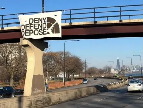 Large white banner reading 'DENY DEFEND DEPOSE' in black lettering, hung from a pedestrian overpass above a roadway.