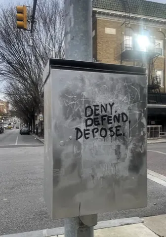 Black marker or paint on a metal utility cabinet reading 'DENY. DEFEND. DEPOSE.' at a city intersection.