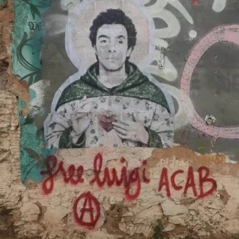 Pasted portrait of a hooded young man on a graffiti-covered wall, with red writing below reading 'free luigi,' 'ACAB,' and a circled-A symbol.