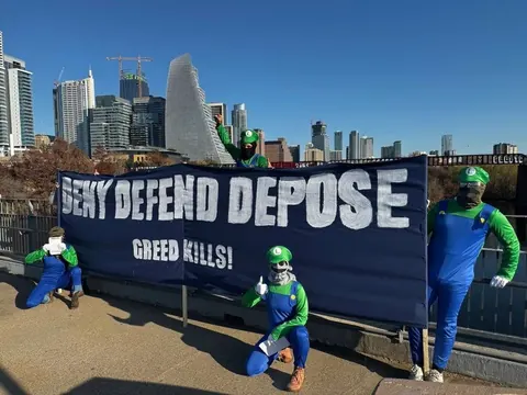 Several people dressed as Luigi pose around a large black banner reading 'DENY DEFEND DEPOSE' with smaller text 'GREED KILLS!' against a city skyline.