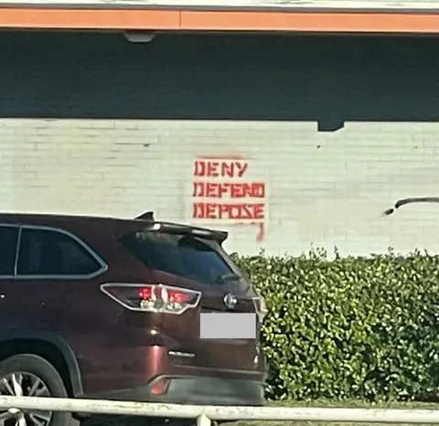 Red stencil-style text reading 'DENY DEFEND DEPOSE' in three stacked lines on a pale brick wall behind a parked vehicle.