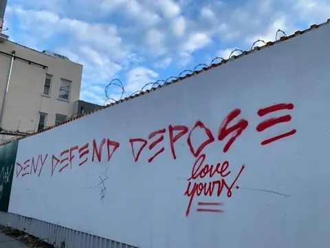 Red graffiti reading 'DENY DEFEND DEPOSE' across a long white wall, with 'love yours' written beneath the larger text.