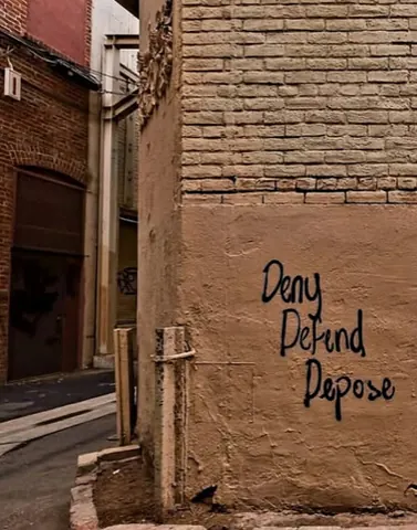 Black handwritten graffiti reading 'Deny Defend Depose' in three stacked lines on a tan alley wall.