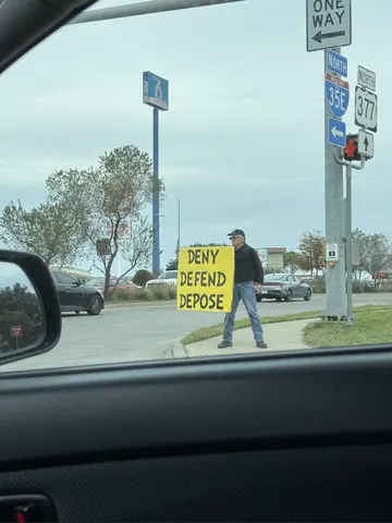Photo taken from inside a vehicle showing a person on a roadside holding a yellow sign reading 'DENY DEFEND DEPOSE.'