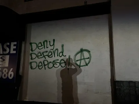Dark graffiti reading 'Delay Defend Depose' beside a green circled-A symbol on a pale wall at night.
