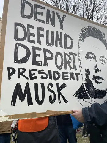 Handheld protest sign reading 'DENY DEFUND DEPORT PRESIDENT MUSK' beside a black-and-white portrait drawing of Elon Musk.
