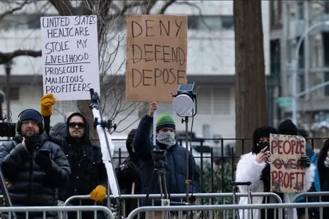 Several protesters behind barricades holding signs, including a central cardboard sign reading 'DENY DEFEND DEPOSE.'