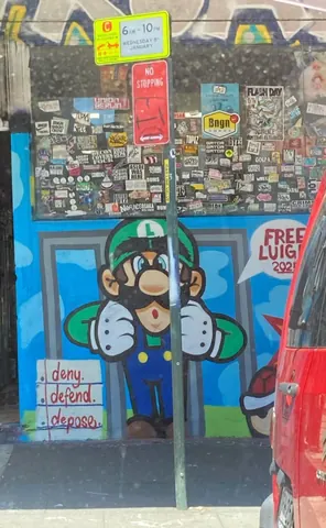 Street mural of Luigi from the Mario games on a sticker-covered storefront or wall, with small 'deny defend depose' text at lower left and 'FREE LUIGI' at right.