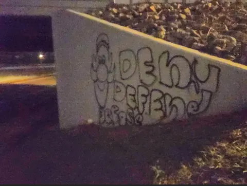 Graffiti on a low concrete wall reading 'DENY DEFEND DEPOSE' in outlined bubble lettering, photographed at night.