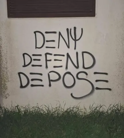 Black graffiti reading 'DENY DEFEND DEPOSE' in three stacked lines on a pale exterior wall above grass.