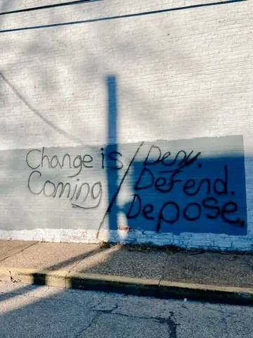 Graffiti on a pale wall reading 'Change is Coming' beside 'Deny Defend Depose,' with dark blue paint and a vertical shadow across the scene.