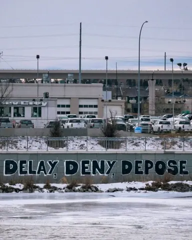 Large black lettering reading 'DELAY DENY DEPOSE' painted across a low roadside barrier in a snowy urban scene.