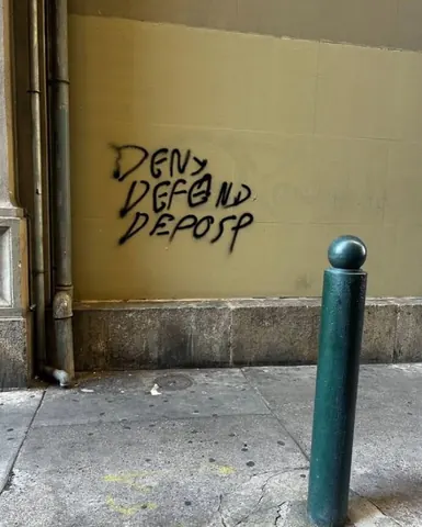 Black graffiti reading 'DENY DEFEND DEPOSE' in three stacked lines on a tan wall beside a sidewalk bollard.