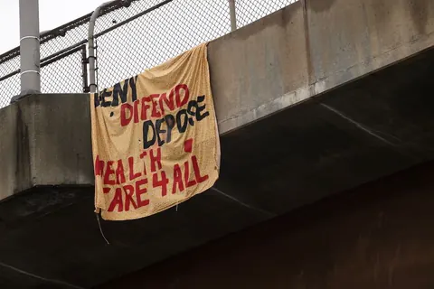 Handmade cloth banner hanging from an overpass with red and black painted text including 'DEFEND,' 'DEPOSE,' and 'HEALTHCARE 4 ALL.'