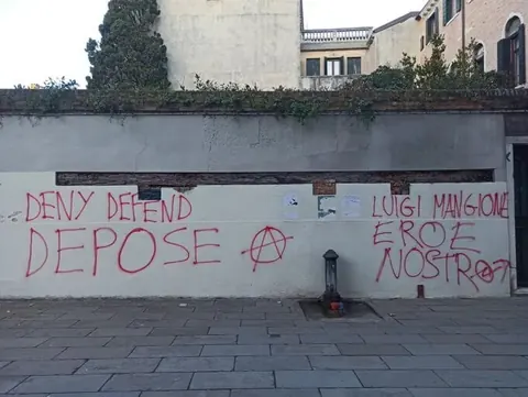 Red graffiti on a long white wall reading 'DENY DEFEND DEPOSE' beside a circled-A symbol, with additional Italian text farther right.