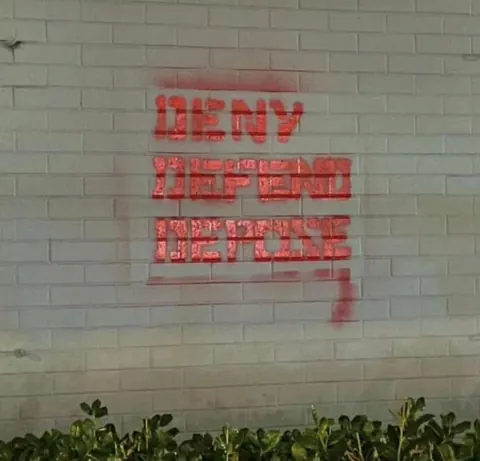 Red stencil-style spray paint reading 'DENY DEFEND DEPOSE' in three stacked lines on a light-colored brick wall.