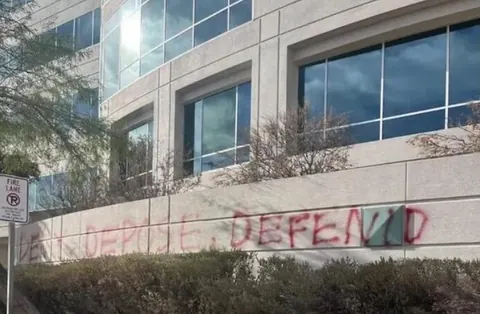 Faded red graffiti reading 'DENY DEFEND' across a low exterior wall in front of a modern building.