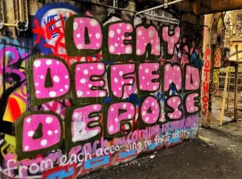 Large colorful graffiti mural with pink bubble letters spelling 'DENY DEFEND DEPOSE' across a heavily tagged wall.