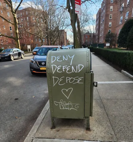 White handwritten text reading 'DENY DEFEND DEPOSE' on a gray street utility box, with a heart and small tag beneath.