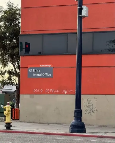 Small pale graffiti reading 'DENY DEFEND DEPOSE' across the lower wall of an orange-and-gray building near an 'Entry Rental Office' sign.