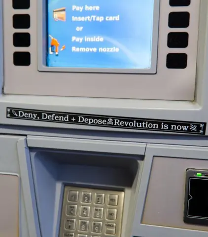 Narrow black sticker placed beneath a payment terminal screen reading 'Deny, Defend + Depose' followed by 'Revolution is now.'