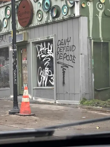 Black graffiti reading 'DENY DEFEND DEPOSE' on the corner wall of a tagged commercial building.