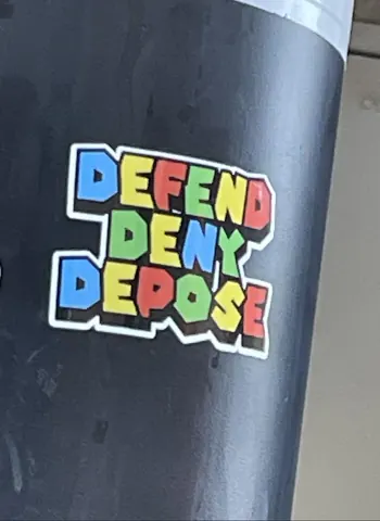 Colorful sticker on a dark pole reading 'DEFEND DENY DEPOSE' in chunky multicolored letters.