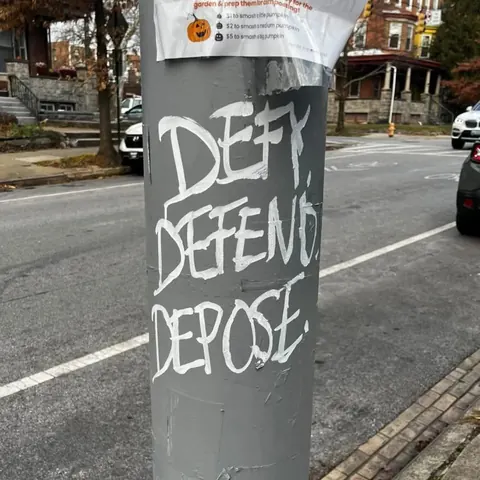 White graffiti reading 'DENY DEFEND DEPOSE' painted vertically on a gray utility pole beside a street.