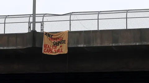 A small handmade banner hanging from an overpass railing, with partially legible red and black text including 'DEPOSE' and 'HEALTHCARE.'