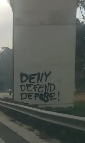 Black spray-painted text reading 'DENY DEFEND DEPOSE!' on a concrete bridge or overpass support beside a roadway guardrail.