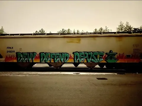 Large green-and-black graffiti lettering reading 'DENY DEFEND DEPOSE' across a long concrete wall beside a roadway.